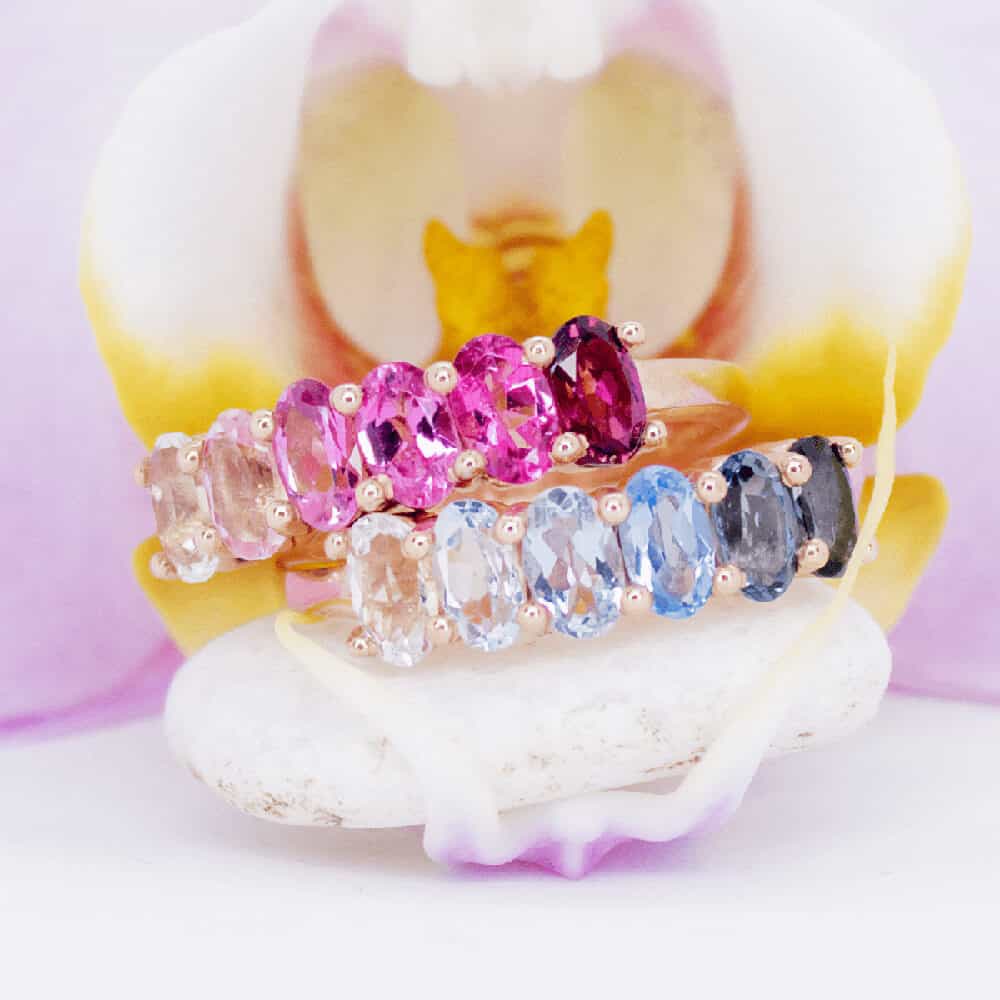 Stunning Ocean Blue and Pink Pomegranate Ombré™ Rings with a strip of six 5 x 3mm oval Topaz gemstones, complemented by a beautiful flower backdrop.