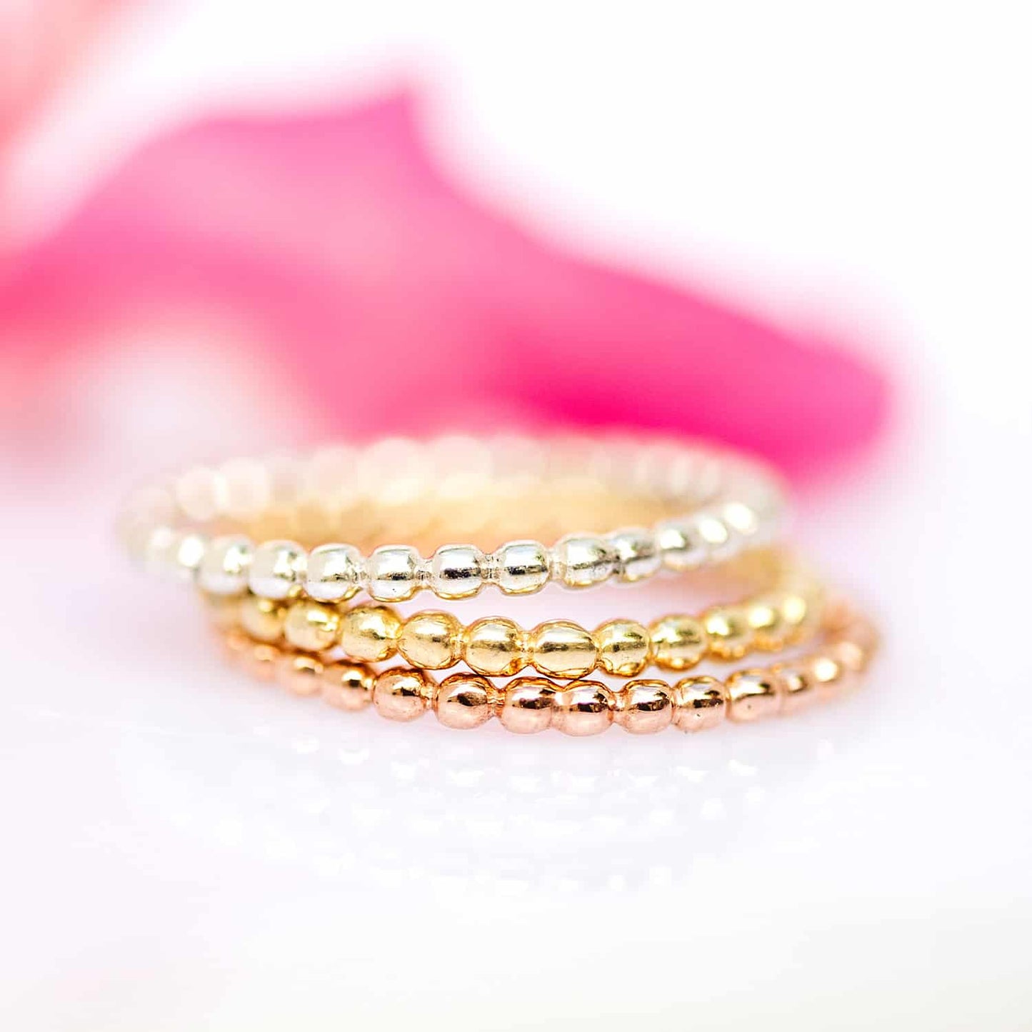 Close-up of stacked Ball rings in sterling silver, yellow, and rose gold plated versions on a white background, accompanied by a pink flower. Versatile and elegant jewellery for any occasion.