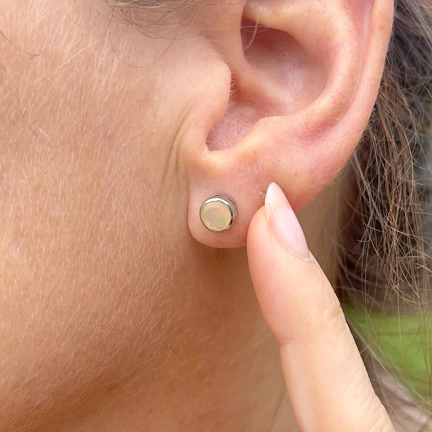 Close-up front view of a woman wearing the Ethiopian Opal Tube Set Studs pointing with her finger at the studs, highlighting a 5mm round Ethiopian Opal gemstone set in sterling silver tube setting.