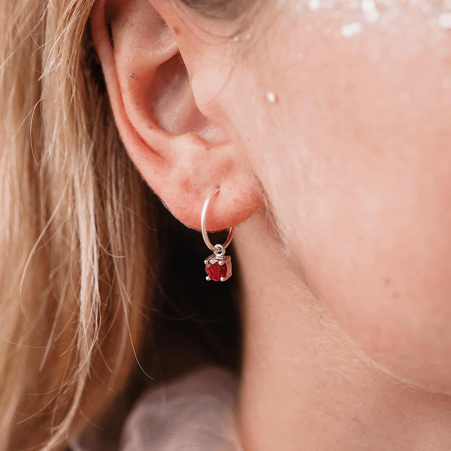 Close up side view of a woman wearing Ruby Dangle Hoop earrings, showcasing a detachable 5mm round Ruby gemstone set in four claws on a Sterling Silver hoop.