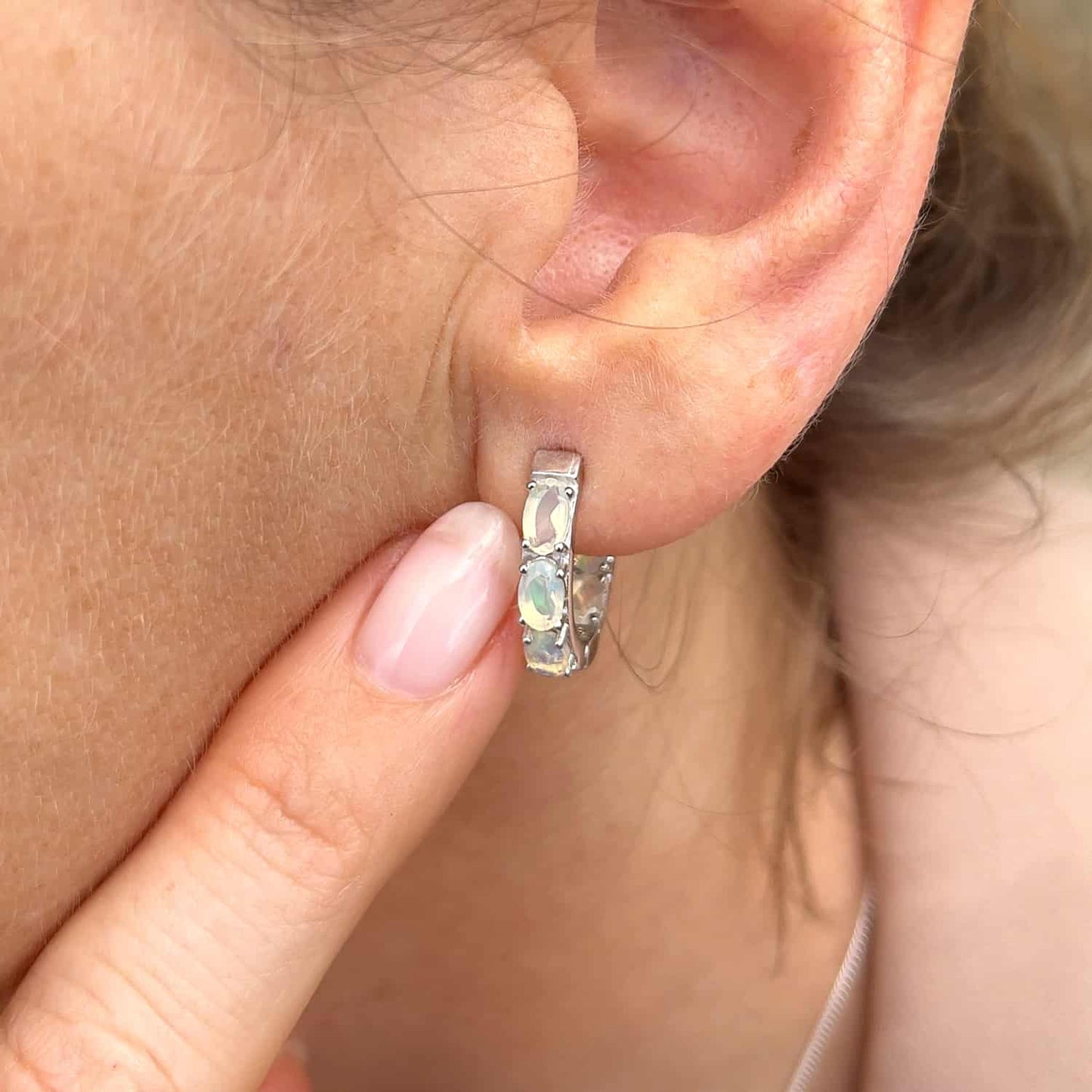 Woman's ear viewed from the front, pointing at Ethiopian Opal Huggie earring. Features six oval 5 x 3mm Ethiopian Opal gems set in Sterling Silver huggie hoops.