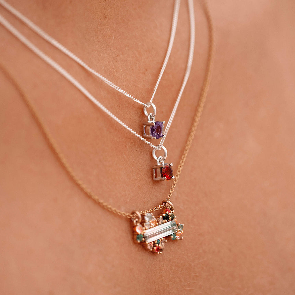 Close-up of a woman wearing the Ruby Pendant necklace with a 5mm round Ruby gemstone set in four claws on a 45cm sterling silver curb chain, layered with two other necklaces.