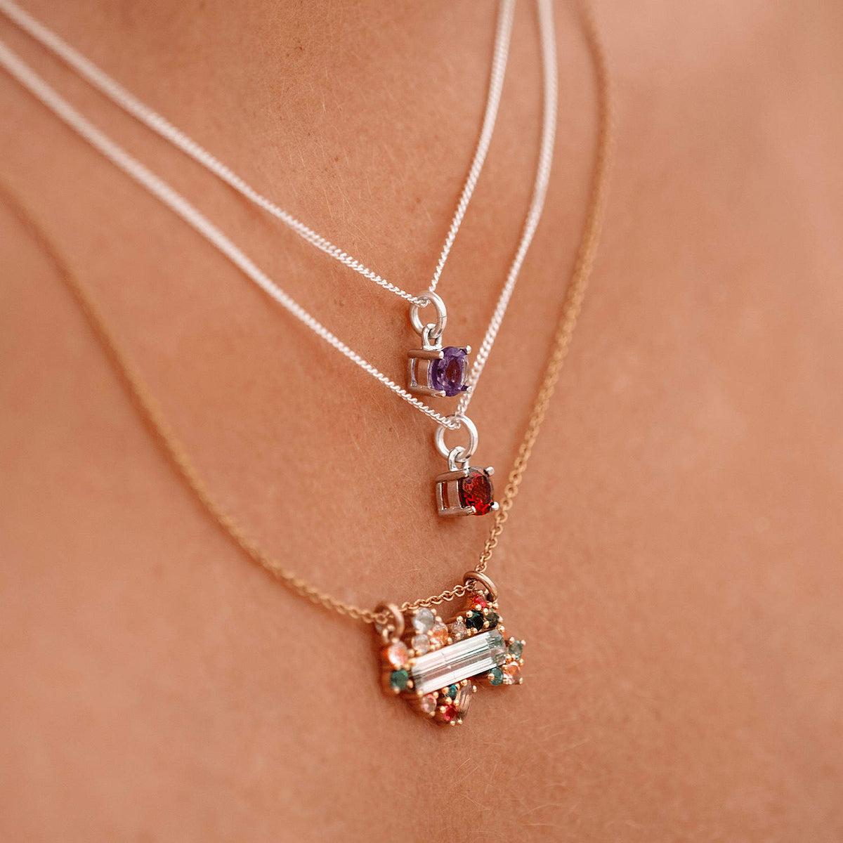 Close-up of a woman wearing the Ruby Pendant necklace with a 5mm round Ruby gemstone set in four claws on a 45cm sterling silver curb chain, layered with two other necklaces.