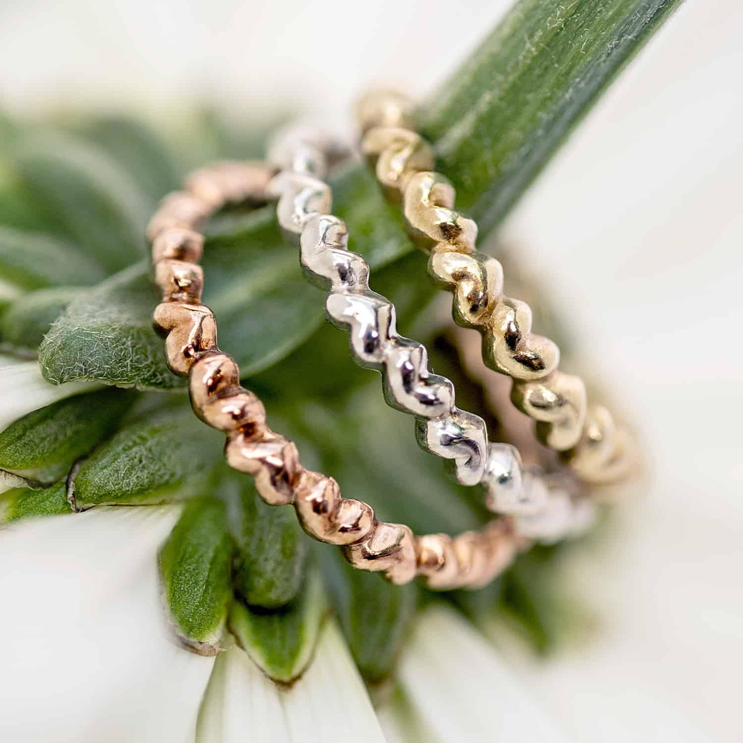 A Sterling Silver heart-shaped ring with multiple interlocking hearts forming a band in silver, yellow gold, and rose gold displayed on a flower