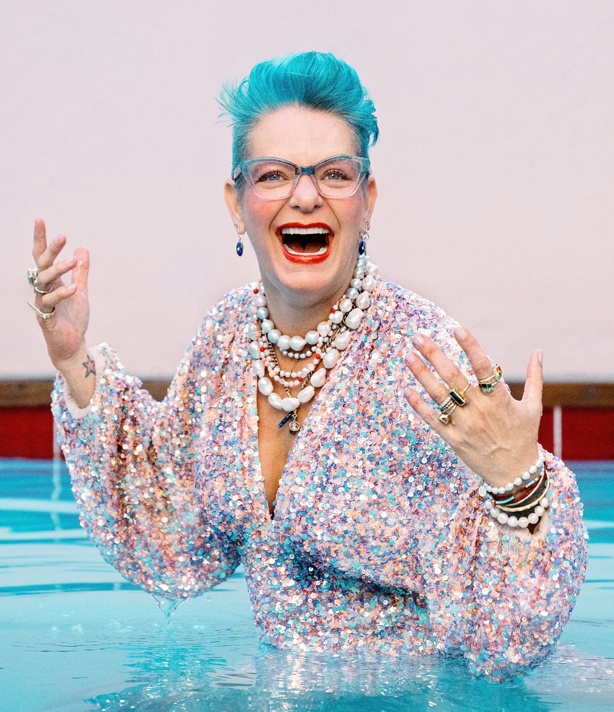 Janine Binneman waist-deep in a swimming pool, wearing a sparkling multicolor long-sleeve top and layered pearl necklaces, with her hands raised in an expressive pose.